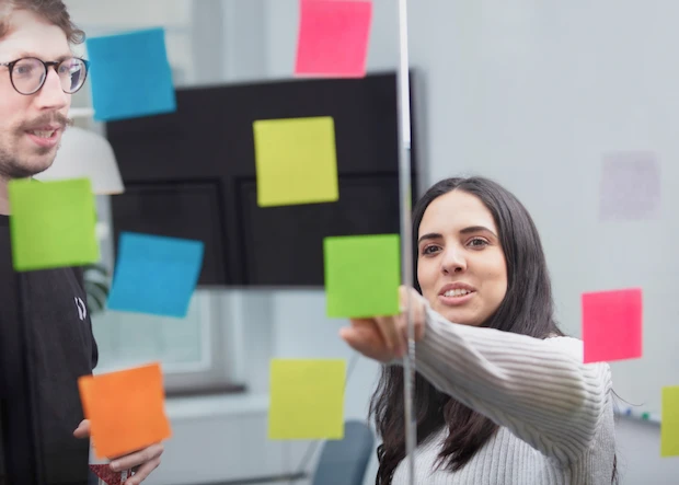 En man och en kvinna står vid en glasvägg där det sitter post-it-lappar i olika färger. kvinnan pekar.