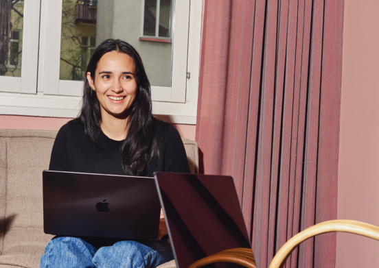 Person med svart tröja arbetar på MacBook i ljust vardagsrum med rosa gardiner och beige soffa, solig och avslappnad kontorsmiljö
