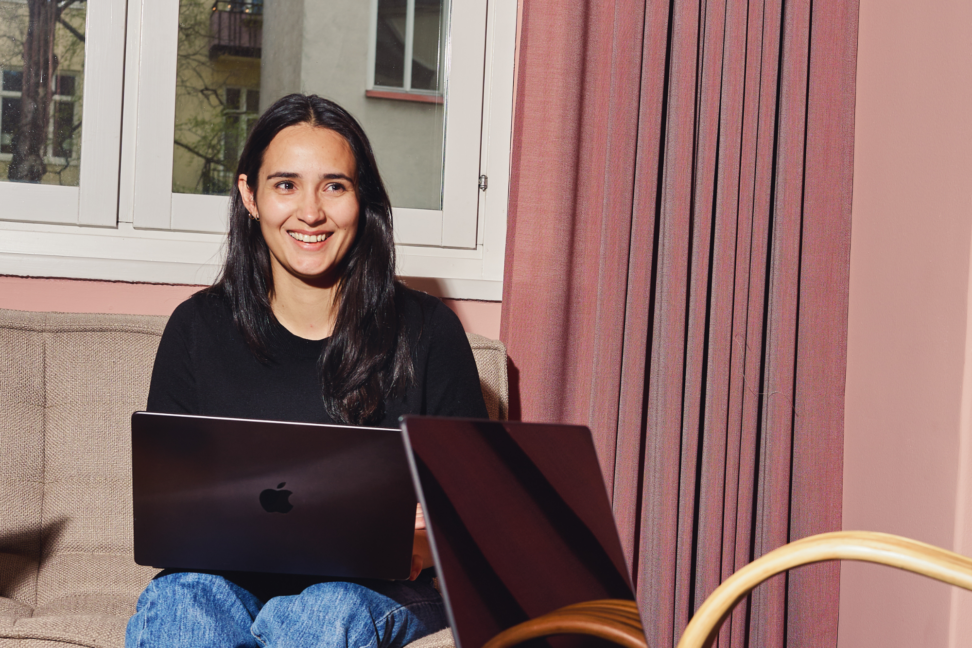 Person med svart tröja arbetar på MacBook i ljust vardagsrum med rosa gardiner och beige soffa, solig och avslappnad kontorsmiljö