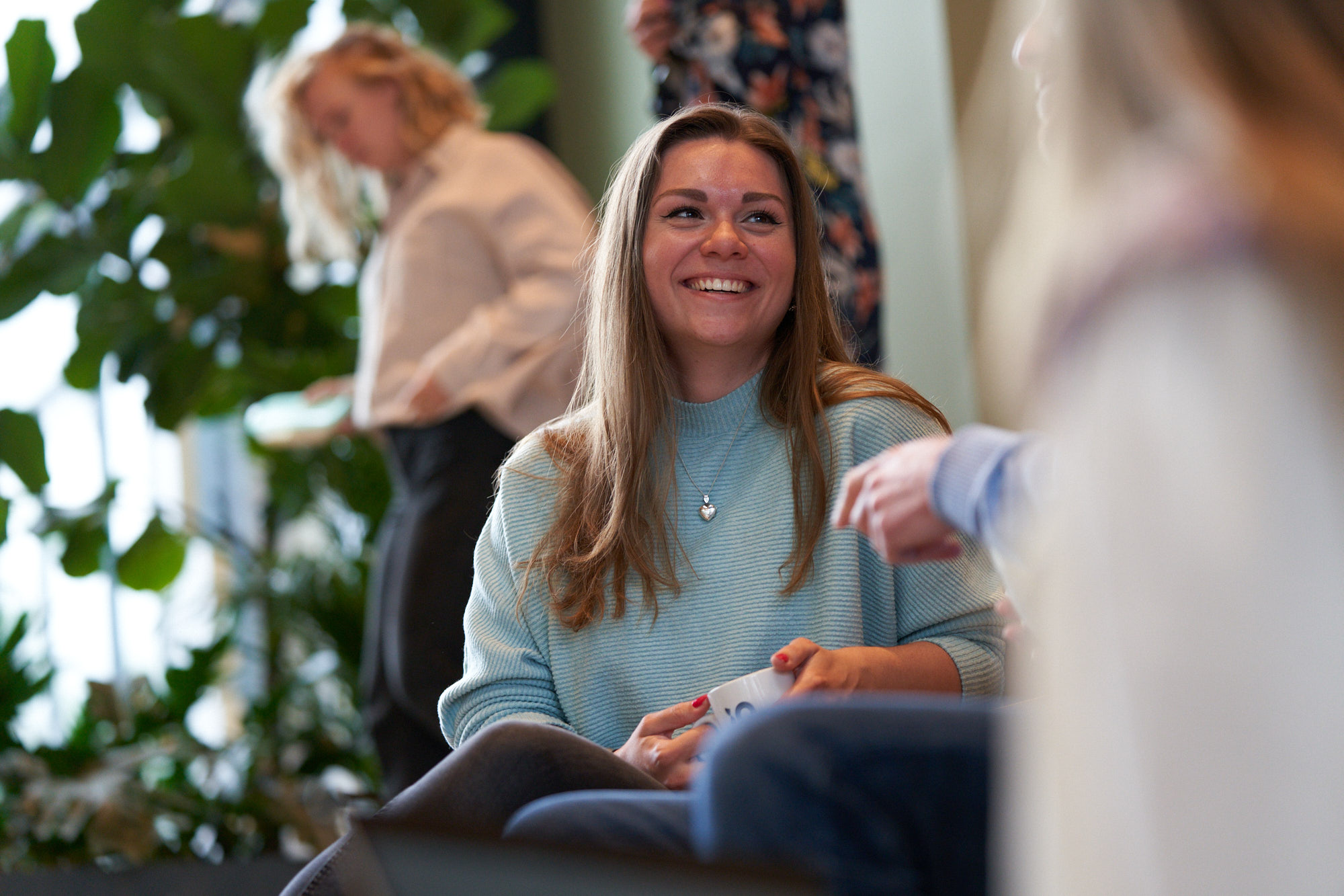 En person i en ljusblå stickad tröja håller en vit mugg. Personen bär ett halsband med ett hjärtformat hänge, och omgivningen inkluderar suddiga silhuetter av andra personer och en grön växt i bakgrunden.
