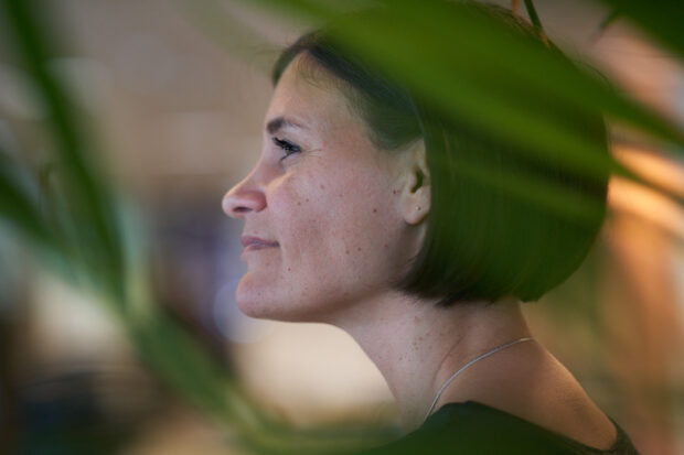 En person med kort mörkt hår ses från sidan, omgiven av gröna växtblad i förgrunden. Bakgrunden är suddig och upplyst.