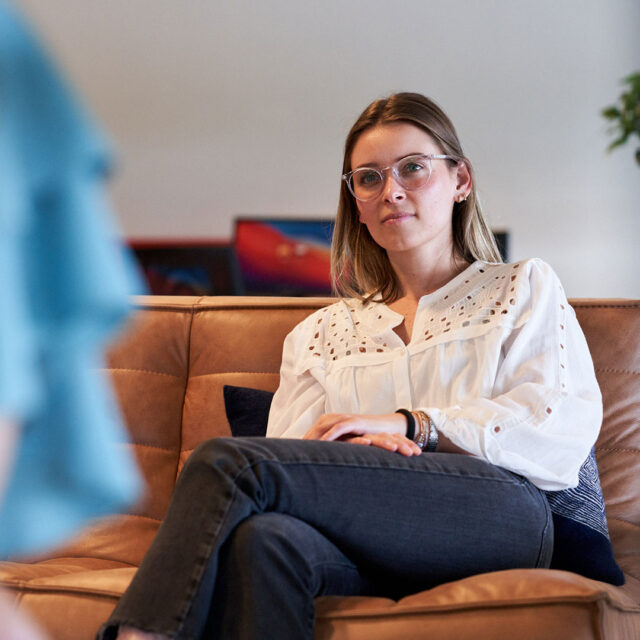 En person sitter på en brun lädersoffa, klädd i en vit blus med broderade detaljer och mörka jeans. Bakgrunden innehåller färgglada tavlor och gröna växter.