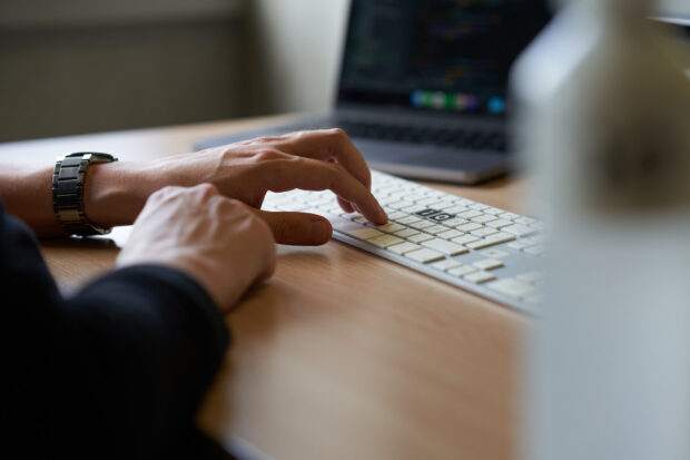 En hand med armbandsur skriver på ett tangentbord på ett skrivbord. En bärbar dator med kod är synlig i bakgrunden.