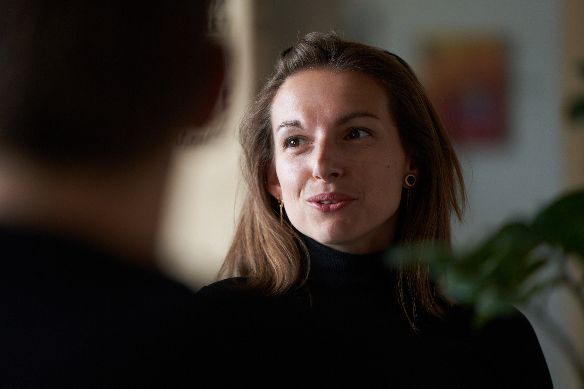 En person med långt brunt hår och svarta kläder står i ett rum med suddig bakgrund, där en växt och en färgglad tavla syns.