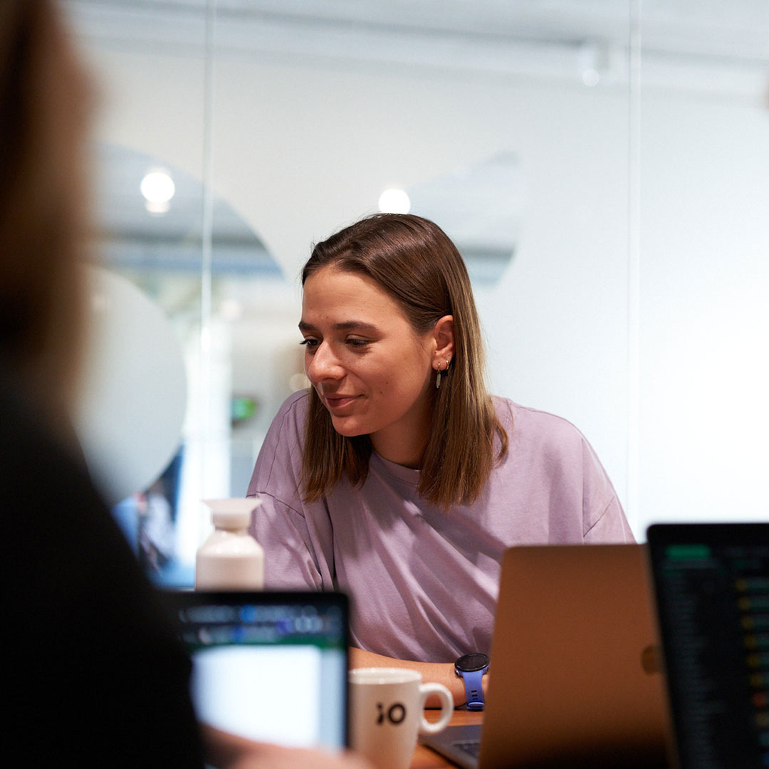 En person med brunt hår och lila tröja sitter vid ett bord med en bärbar dator, en kaffemugg med texten "io", och en vit flaska. Bakgrunden visar en kontorsmiljö med glasväggar.