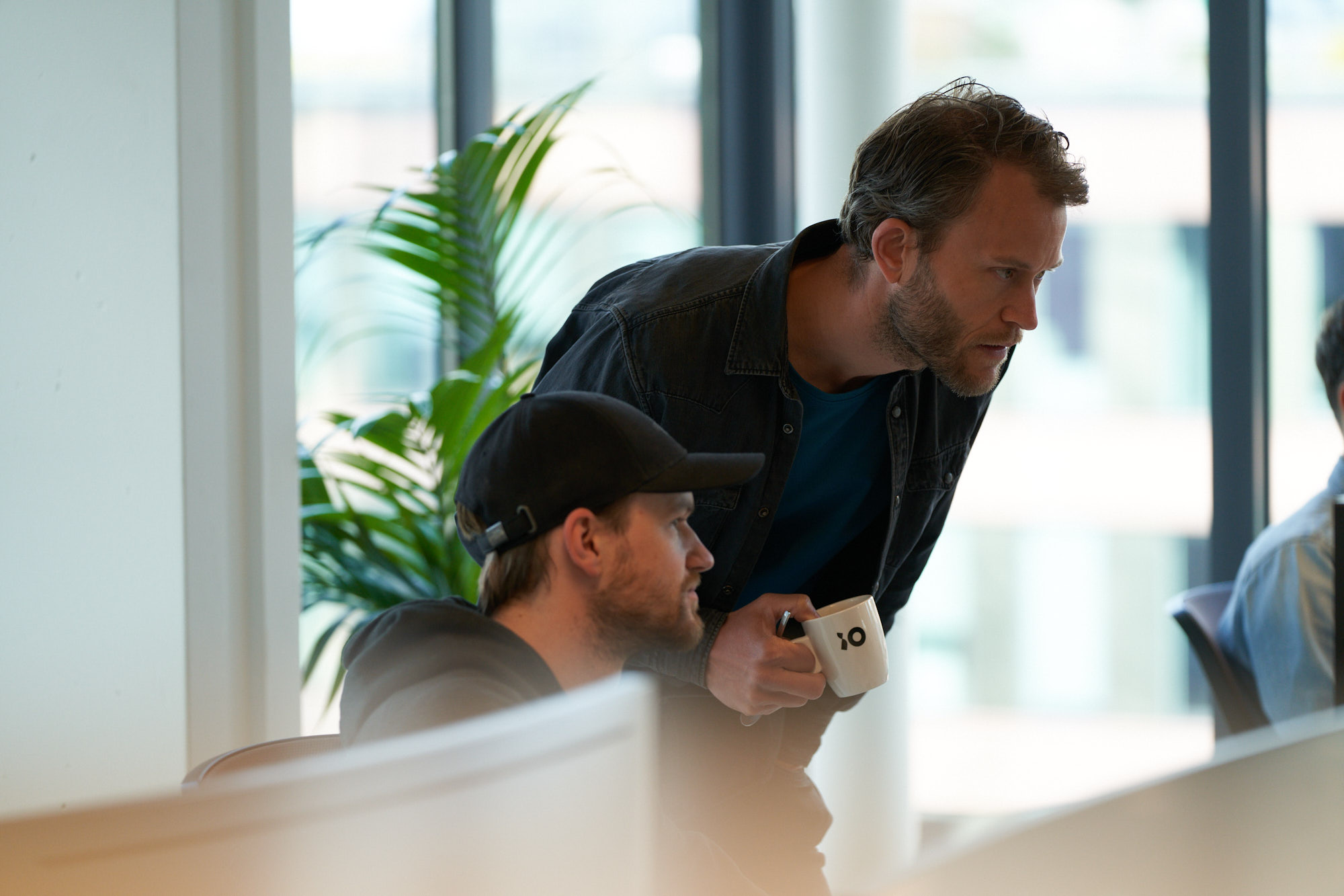 En person håller en vit kaffekopp med en svart symbol och lutar sig över en annan, med en kontorsmiljö i bakgrunden.